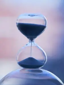 A glass hourglass on a blurred pink and purple background, with dark sand running through from the top to the bottom to represent the passage of time.