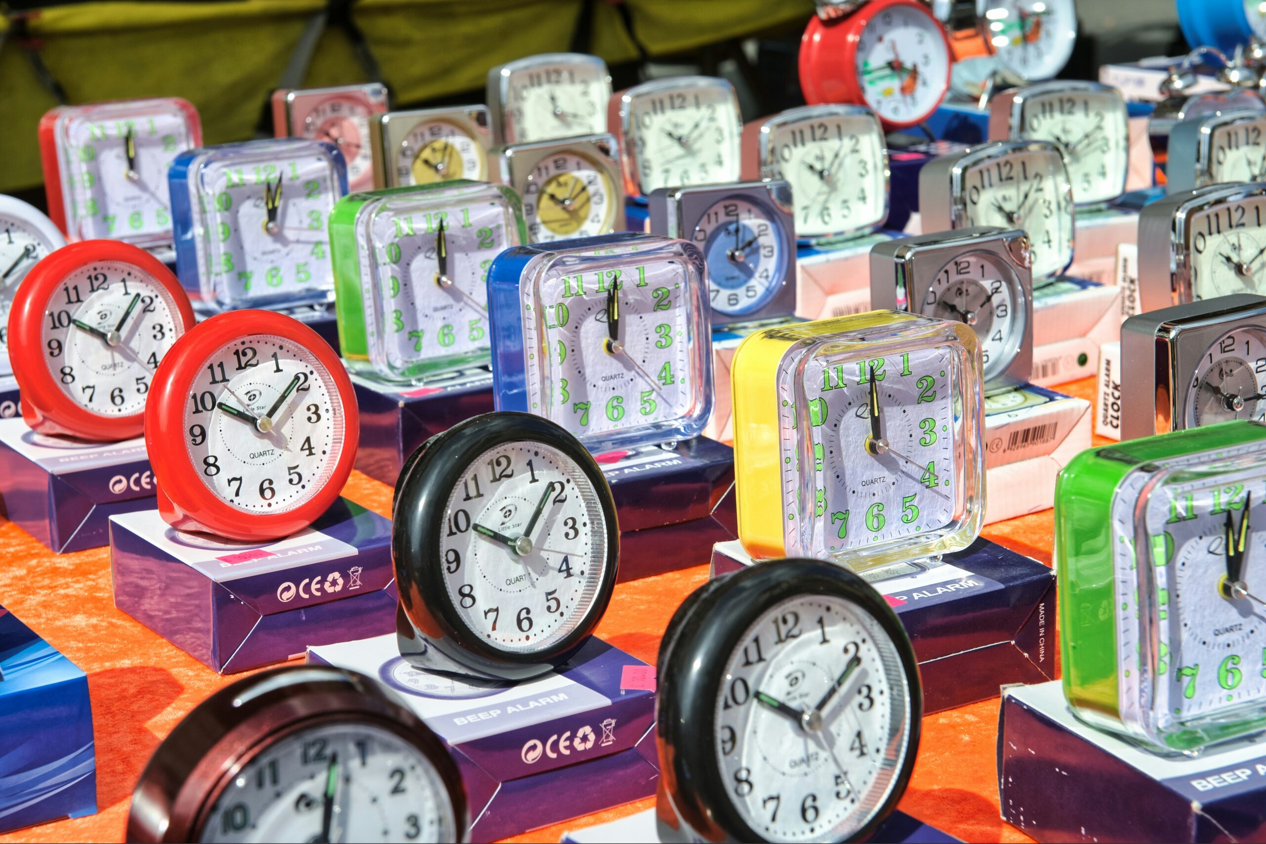 Photo by Alexander Schimmeck of a collection of analogue alarm clocks displayed for sale, featuring both round and square faces in various colours including red, blue, green, yellow, and black. The clocks are arranged on an orange display surface with their retail packaging visible beneath them.