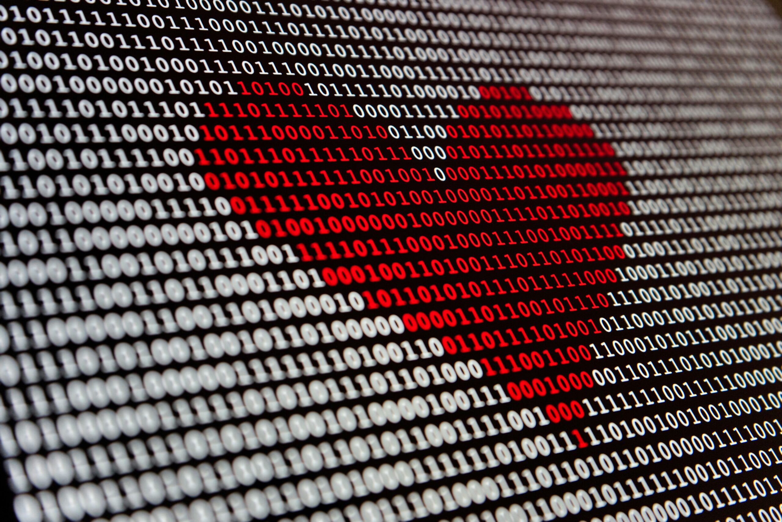 Photo by Alexander Sinn of a close-up of binary code (1s and 0s) displayed on a screen, with some digits highlighted in red forming a pattern against the white digits on a black background. The red digits appear to form a heart shape in the binary pattern.