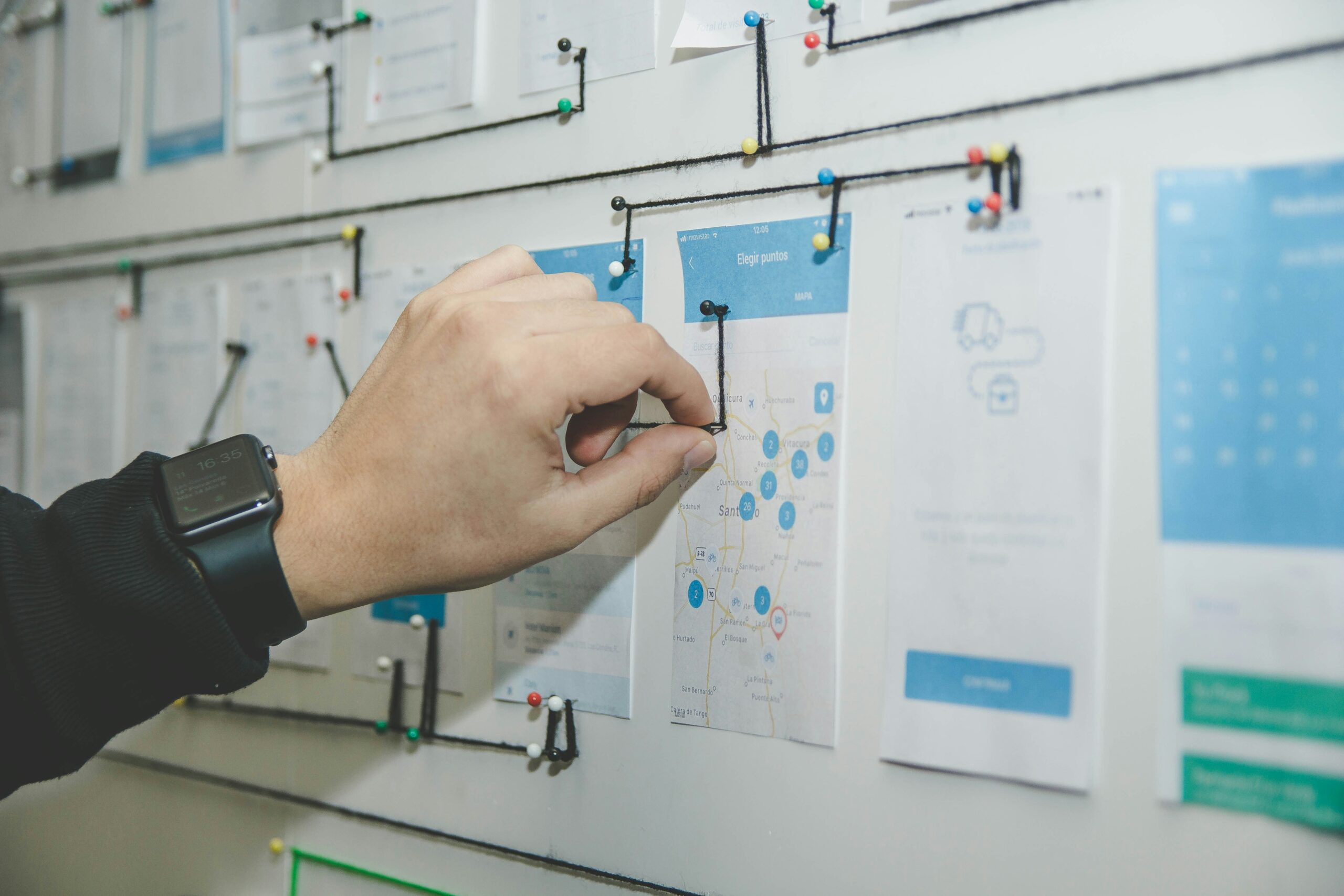 A close-up photo of a person's hand by Alvaro Reyes. The hand is adjusting a pin on a workflow or wireframe board. The board contains multiple sheets of paper displaying app or website design mock-ups, with black string connecting various points. The person is wearing a black smartwatch on their wrist. The background is slightly blurred, emphasizing the detailed planning and organization process.