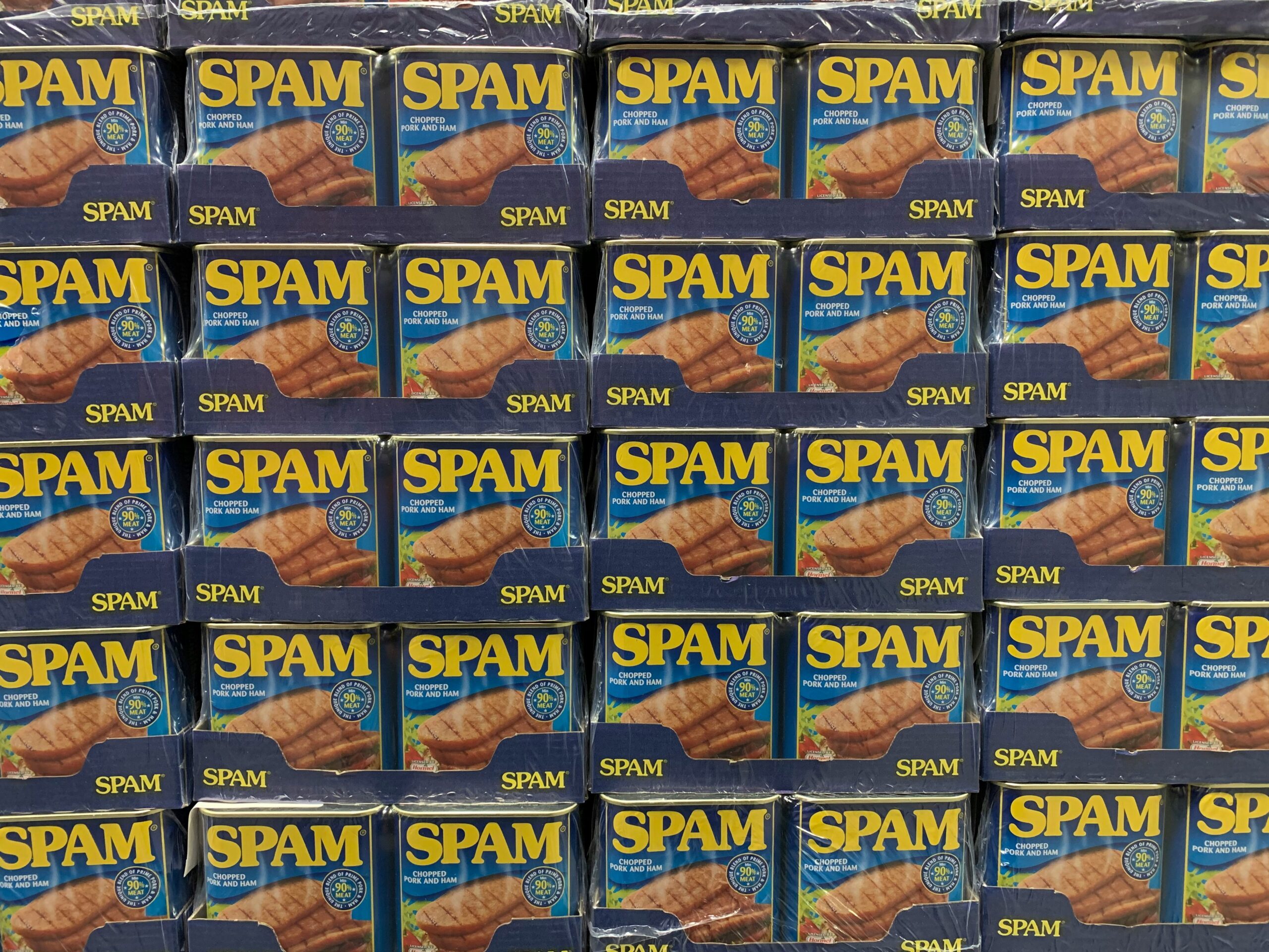 Photo by Hannes Johnson of multiple rows of blue and yellow SPAM canned meat product boxes stacked on store shelves. Each box shows the SPAM logo and indicates it contains 'Chopped Pork and Ham' with a product certification seal.