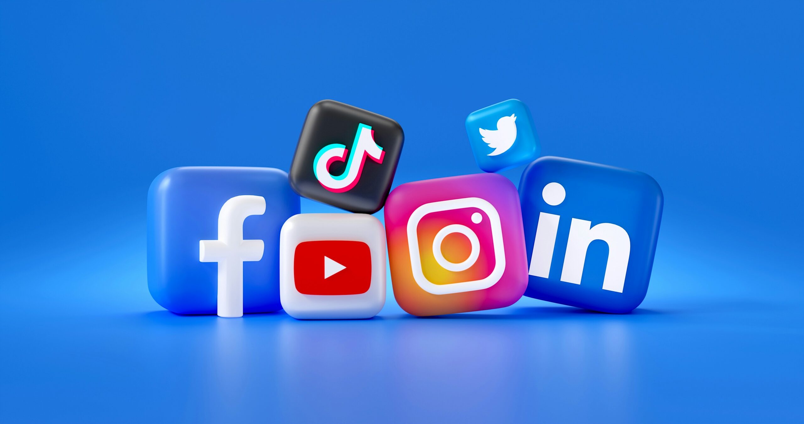 A 3D-rendered digital image by Mariia Shalabaieva of various social media app icons arranged in a playful composition on a blue background. The icons include Facebook, YouTube, Instagram, LinkedIn, Twitter, and TikTok, each represented as rounded cubes with their respective logos.