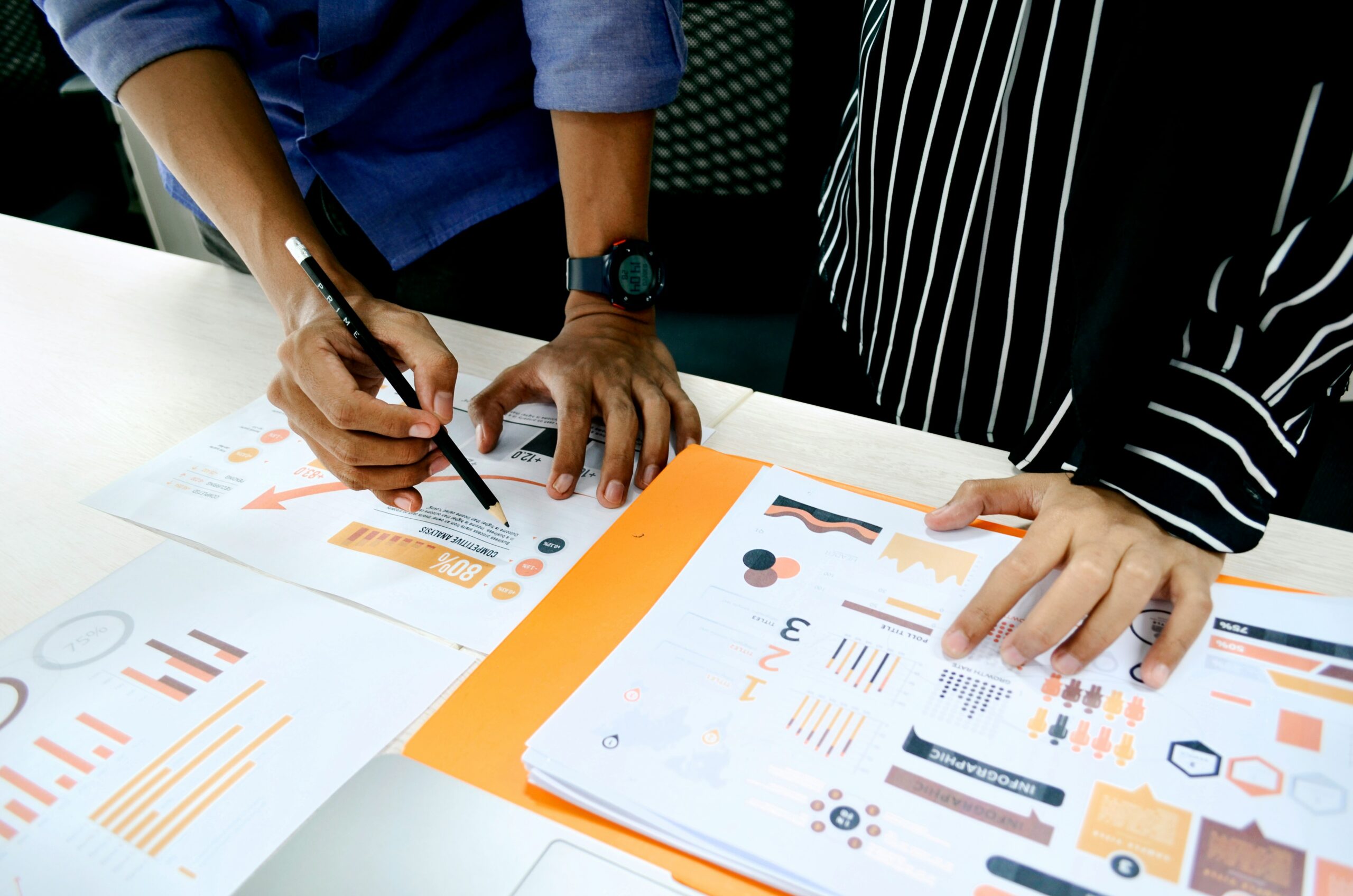 A photo by Ux Indonesia of an overhead view of hands working on business analytics documents spread across a white table. The papers contain orange and black infographics, charts, and data visualizations. One person wearing a blue shirt is writing while another person in a striped garment points at the data.