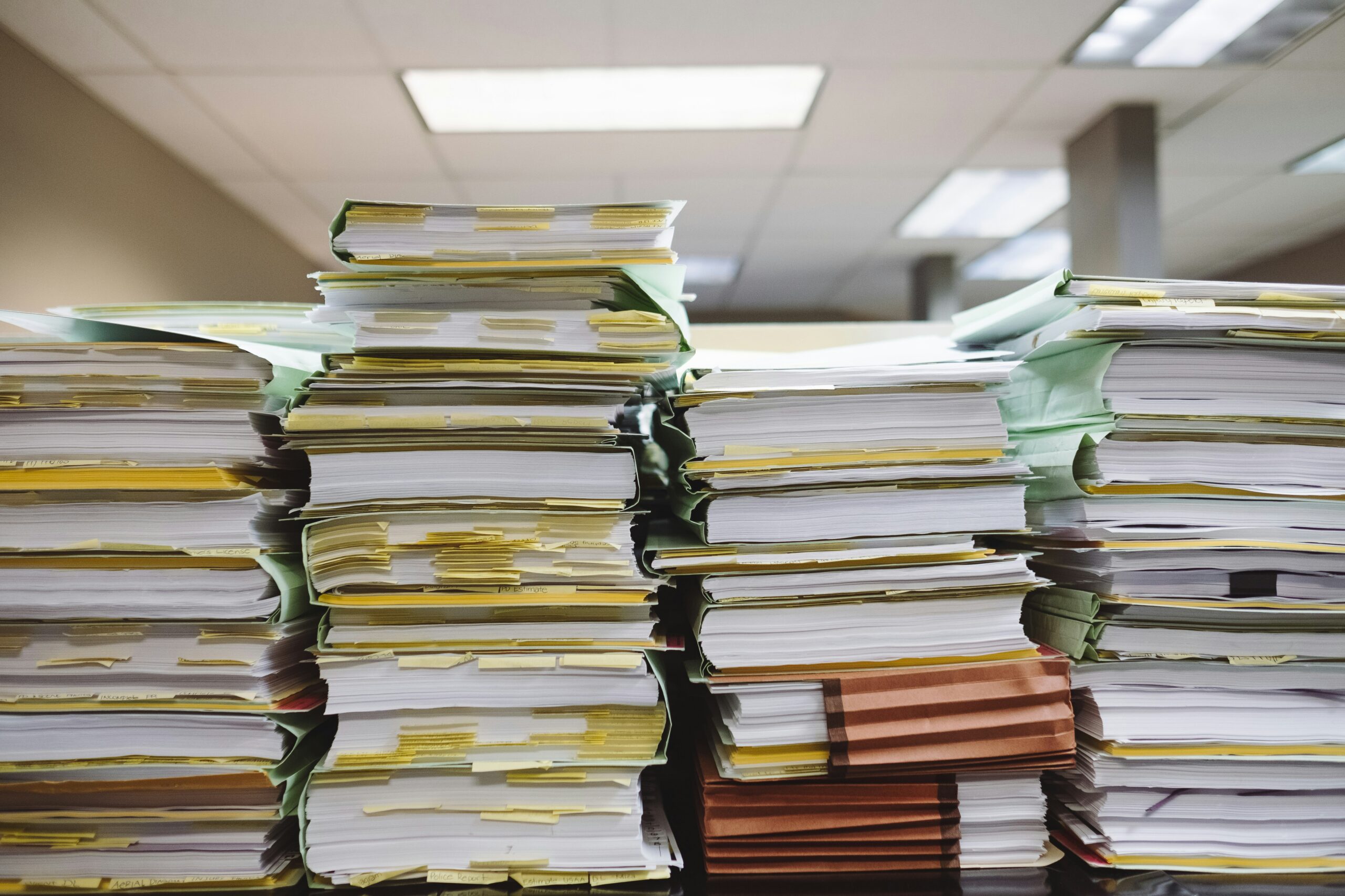 Photo by Wesley Tingey of tall stacks of paper documents and files piled on a surface, with pale green file folders and yellow sticky notes visible throughout. The scene is set in an office environment with fluorescent lighting overhead.